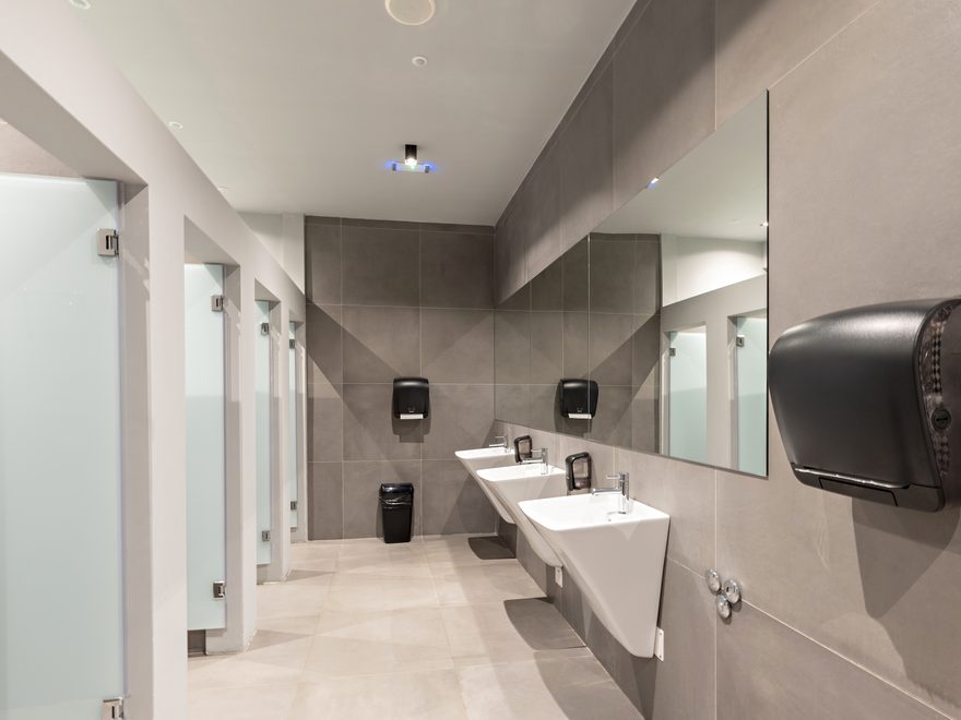 A commercial washroom with grey tiles, a row of wall-hung basins, frosted cubicle doors and a paper towel dispenser