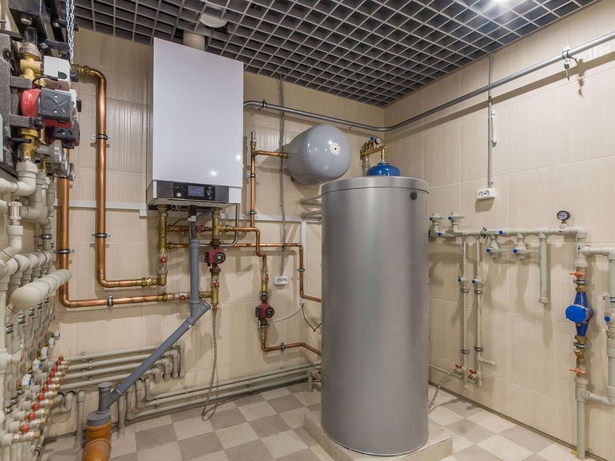 A tiled plant room with a tall silver hot water cylinder, a wall-mounted heater and copper and plastic pipework with valves and gauges