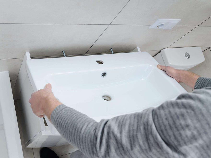 Two hands lifting a white ceramic basin into place on a vanity unit against a tiled wall